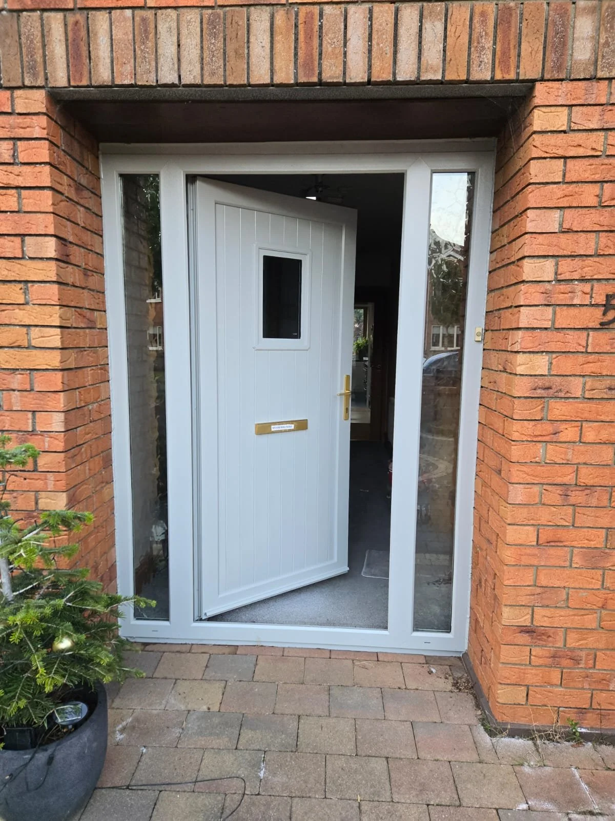 A white front door with a small window and a mail slot, partially open, set in a brick house with glass side panels, and a potted Christmas tree outside.