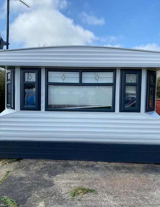 Front view of a white and black mobile home with decorative windows, situated on a paved area under a partly cloudy sky.