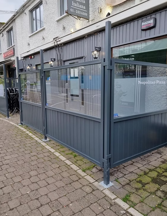 Outdoor seating area enclosed with gray metal and glass panels in front of a restaurant, with cobblestone sidewalk and building behind.