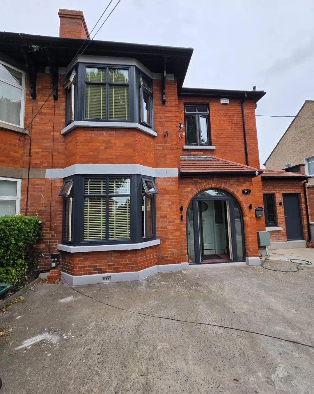 A red brick residential building with bay windows, an arched front door, and a concrete driveway.