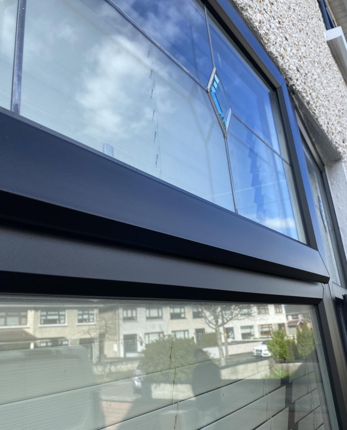 Close-up of a large window with reflective glass, showing the sky with clouds and nearby residential buildings outside, including houses, trees, and a parked car.