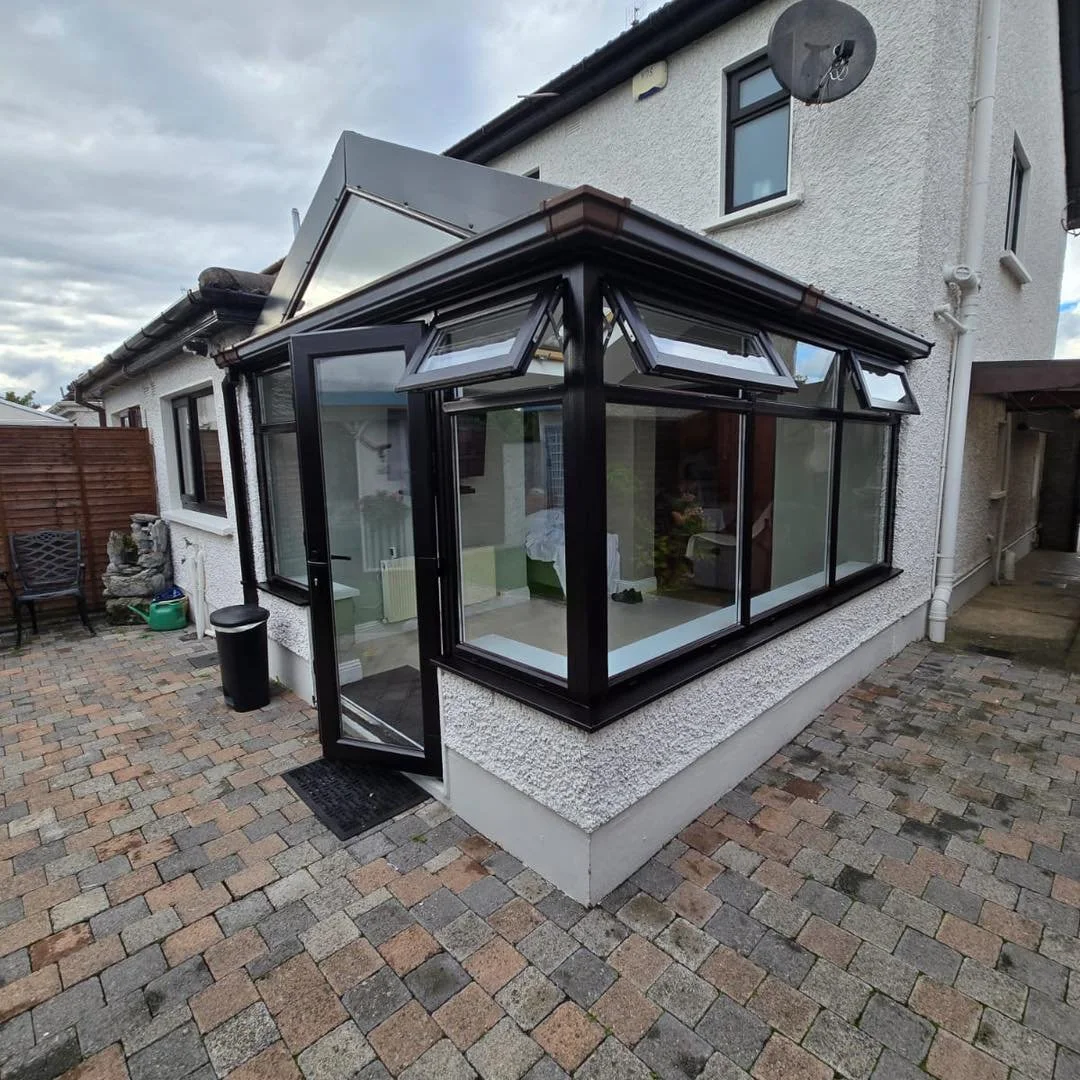 A small enclosed sunroom with black frame windows attached to a white house, with an open door leading outside to a brick patio.