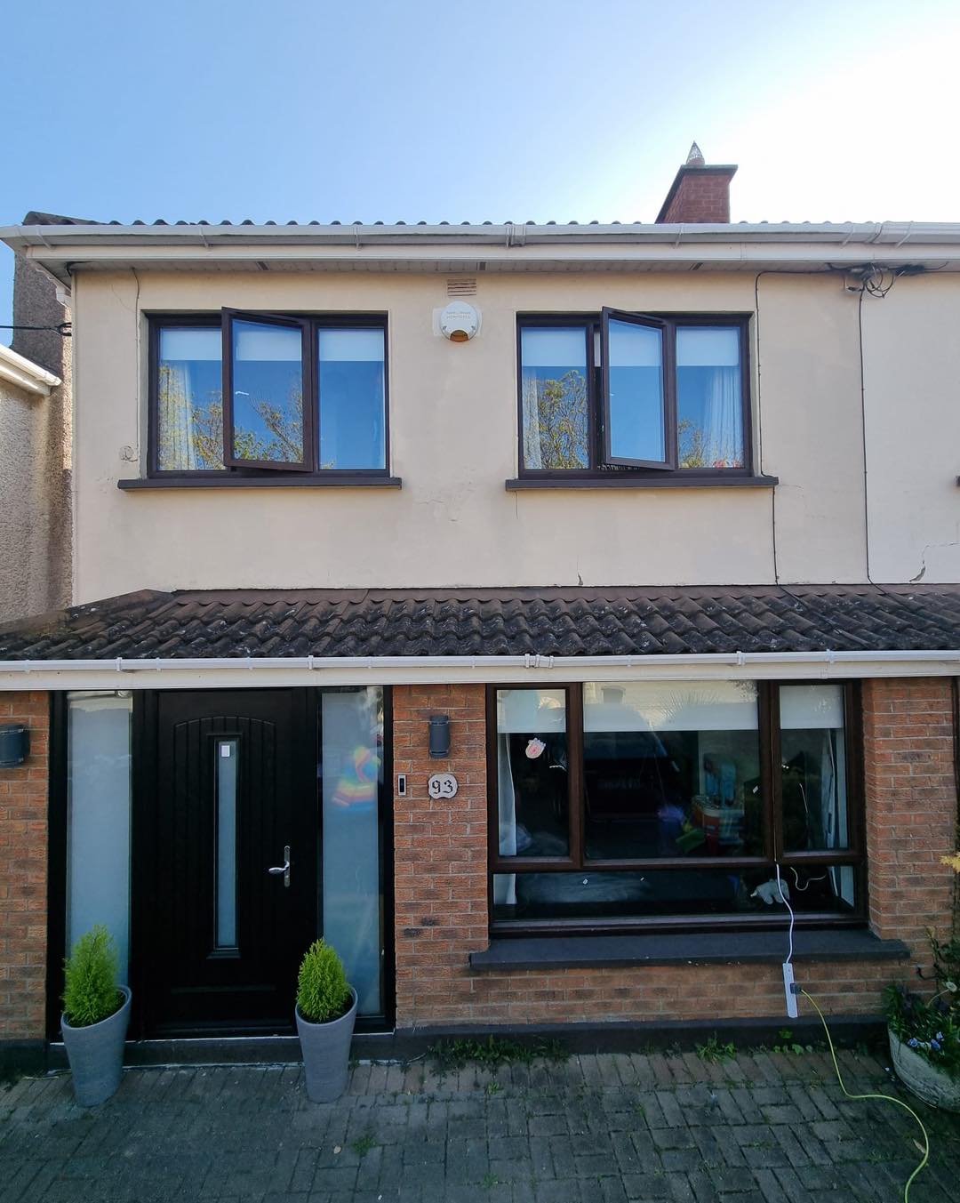 Front view of a two-story house with a brick ground floor, cream upper walls, and a slate roof. The ground floor has a black door with frosted glass and two potted plants on either side. There is a large window to the right of the door with an interi