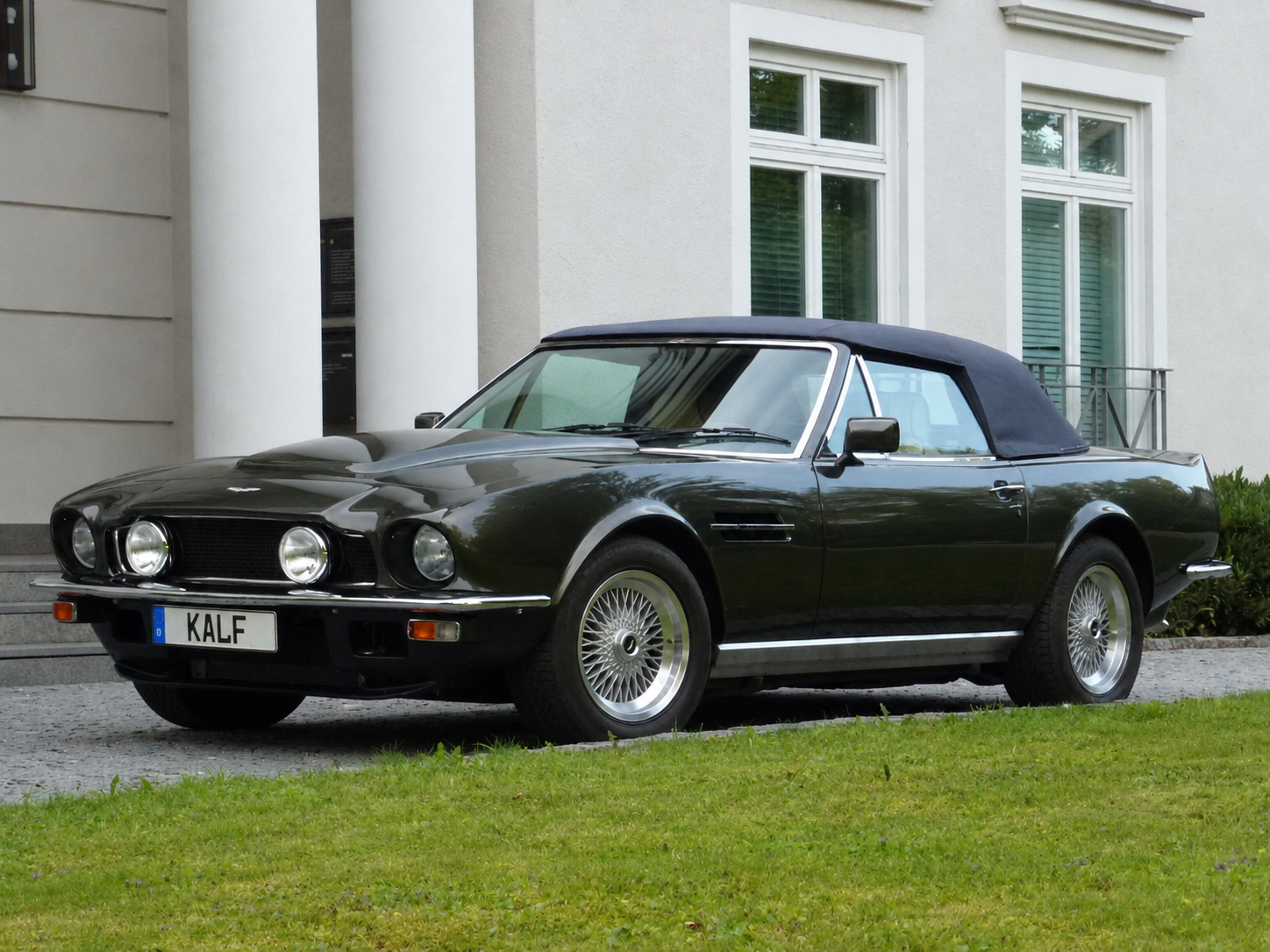 Klassischer grüner Aston Martin vor weißer Villa.png