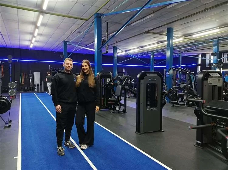 Two people standing in a gym, surrounded by workout equipment, with blue lighting and a blue running track.