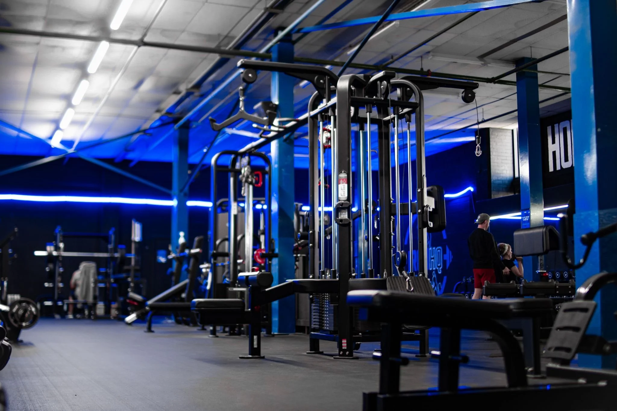 Interior of a modern gym with various workout equipment including weight machines, free weights, and hanging straps, illuminated with blue lighting, with a person standing and another sitting in the background.