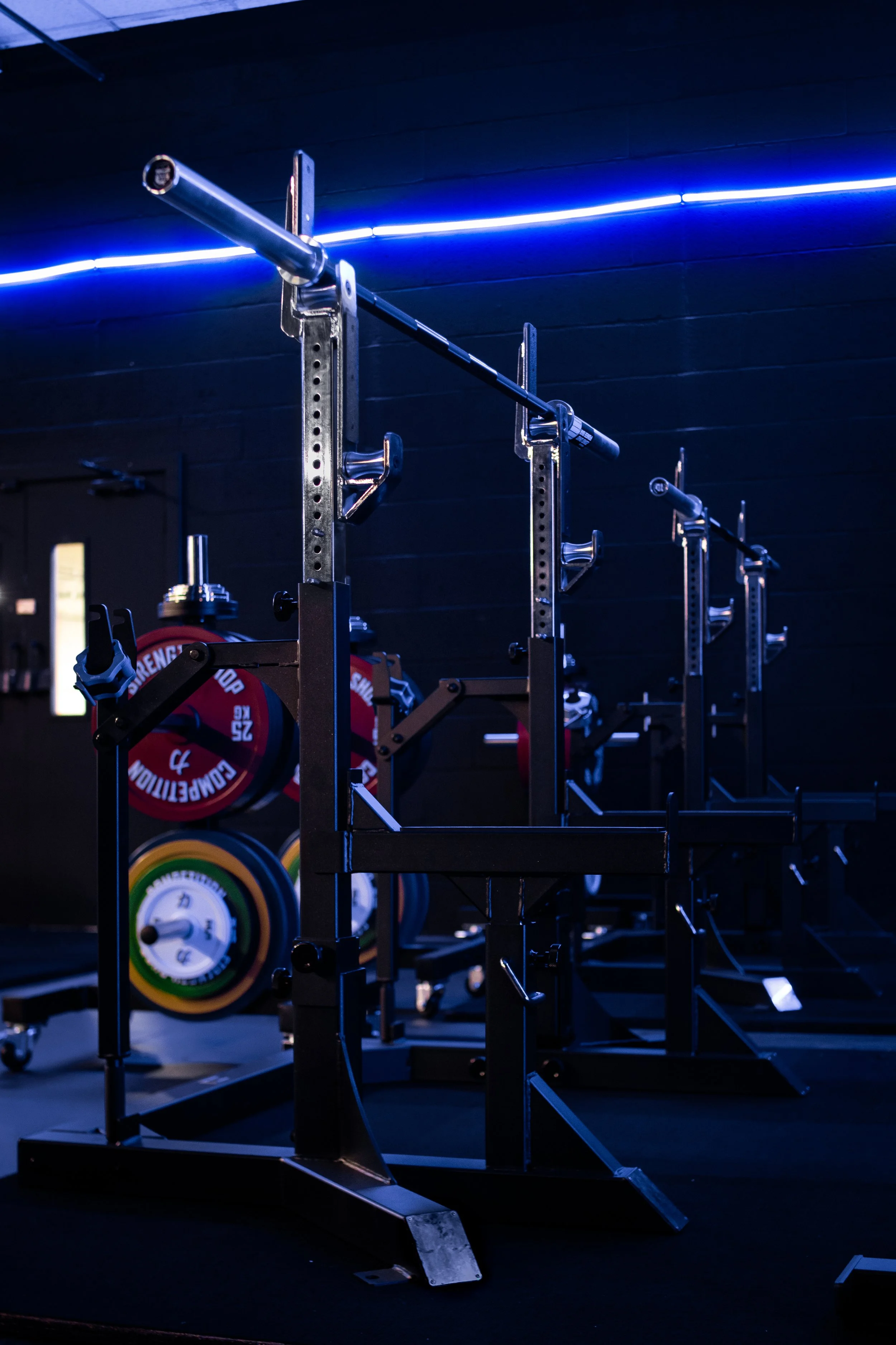 Gym interior with weightlifting squat racks, colorful weight plates, and blue neon lighting