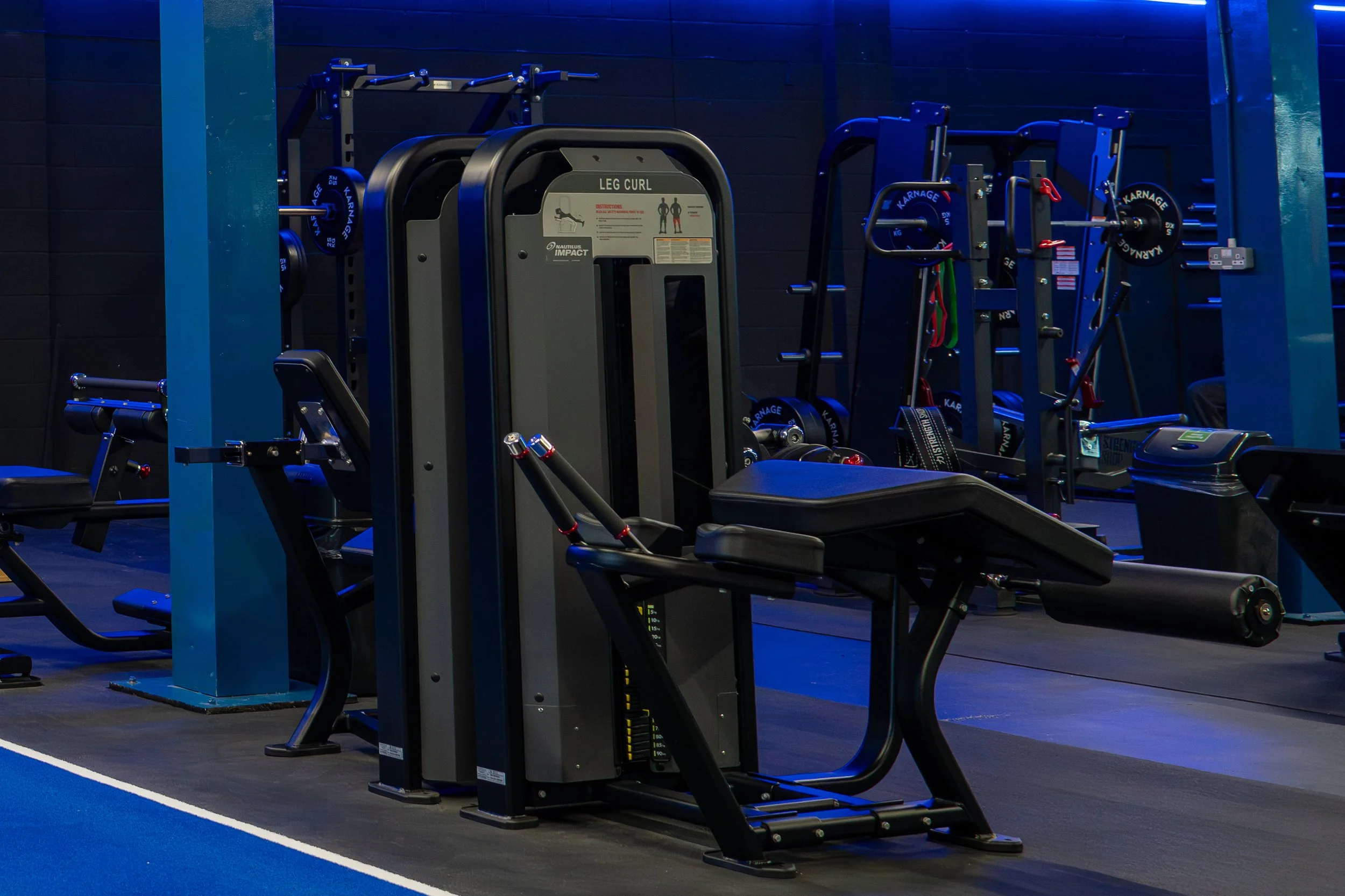 Fitness equipment in a gym, including a leg curl machine, weight plates, resistance bands, and a kettlebell, under blue lighting.