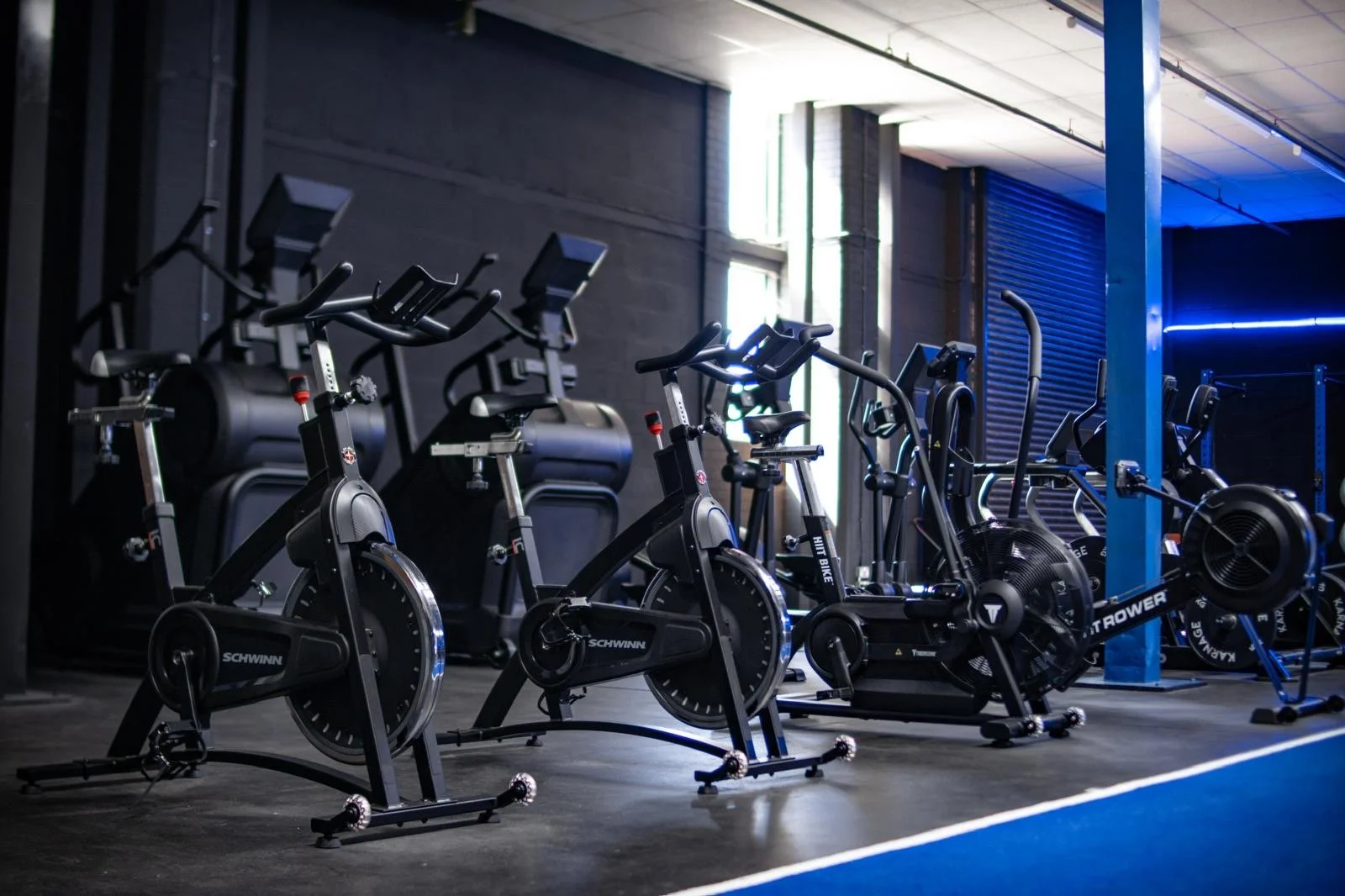 A row of cardio exercise bikes and rowing machines in a modern gym with black walls and blue accent lighting.