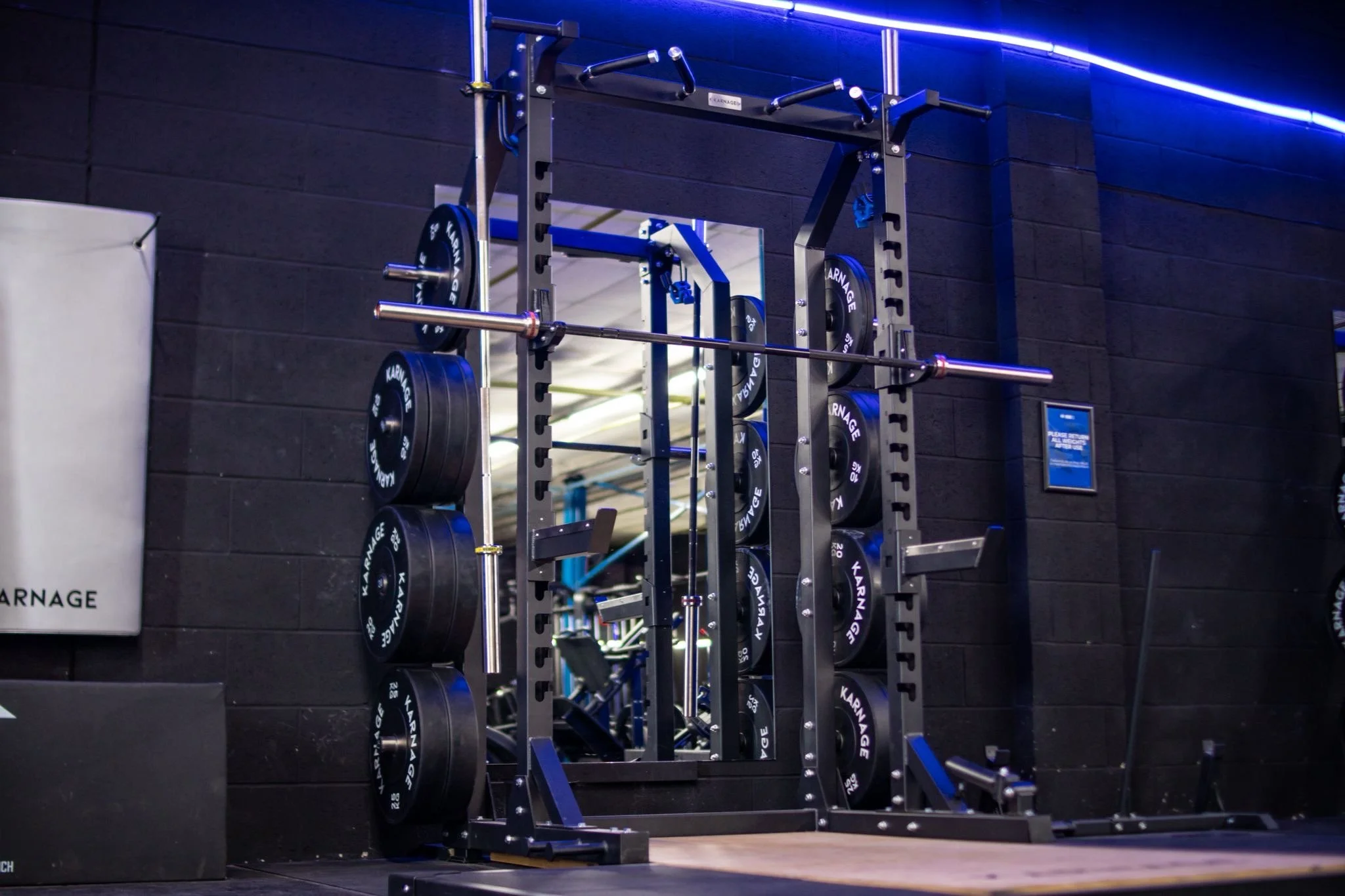 A gym with a black brick wall, blue neon lights, and a workout station with barbells and weight plates.