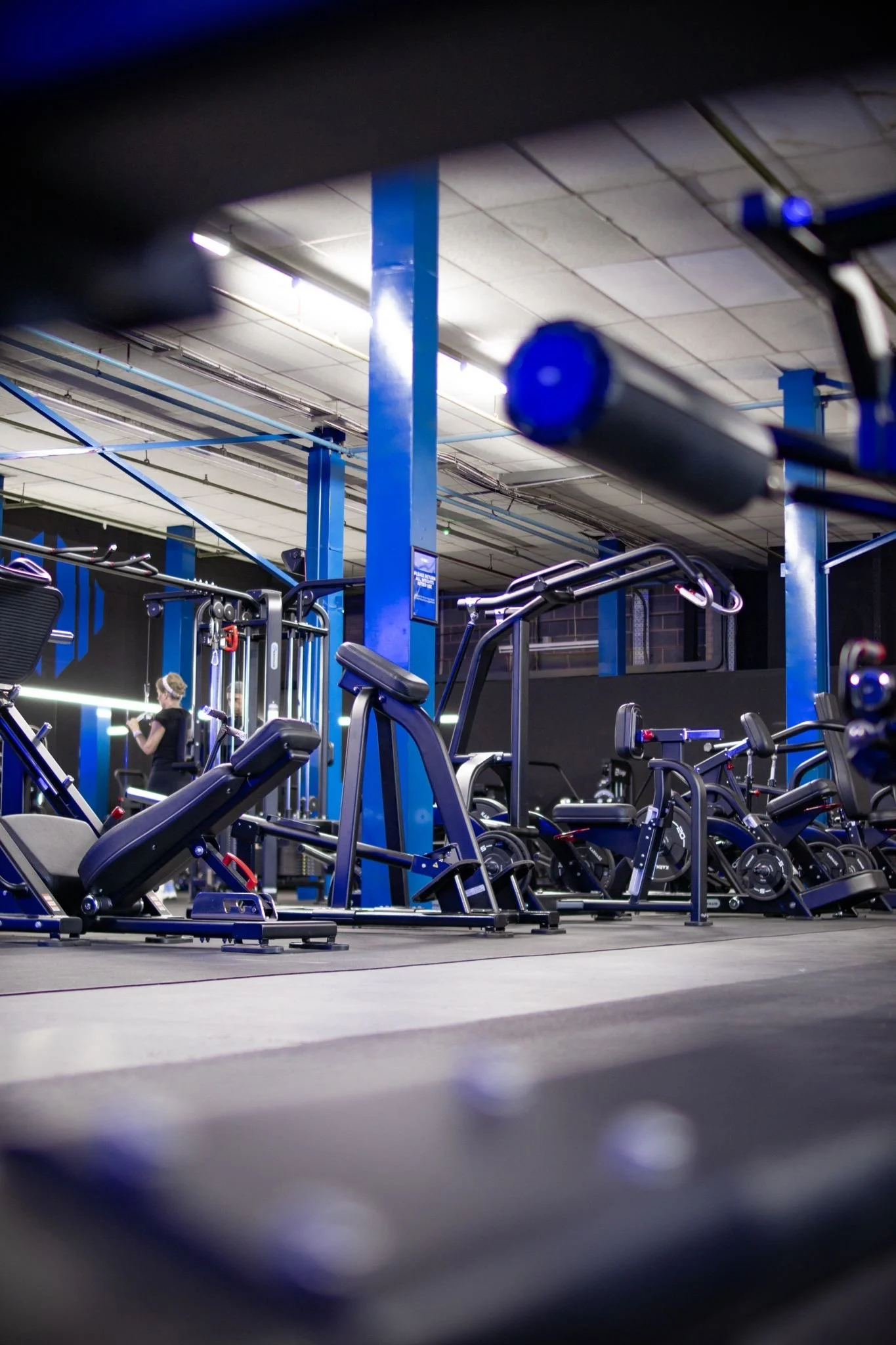 Indoor gym with various workout equipment, including weight machines and a woman walking in the background.