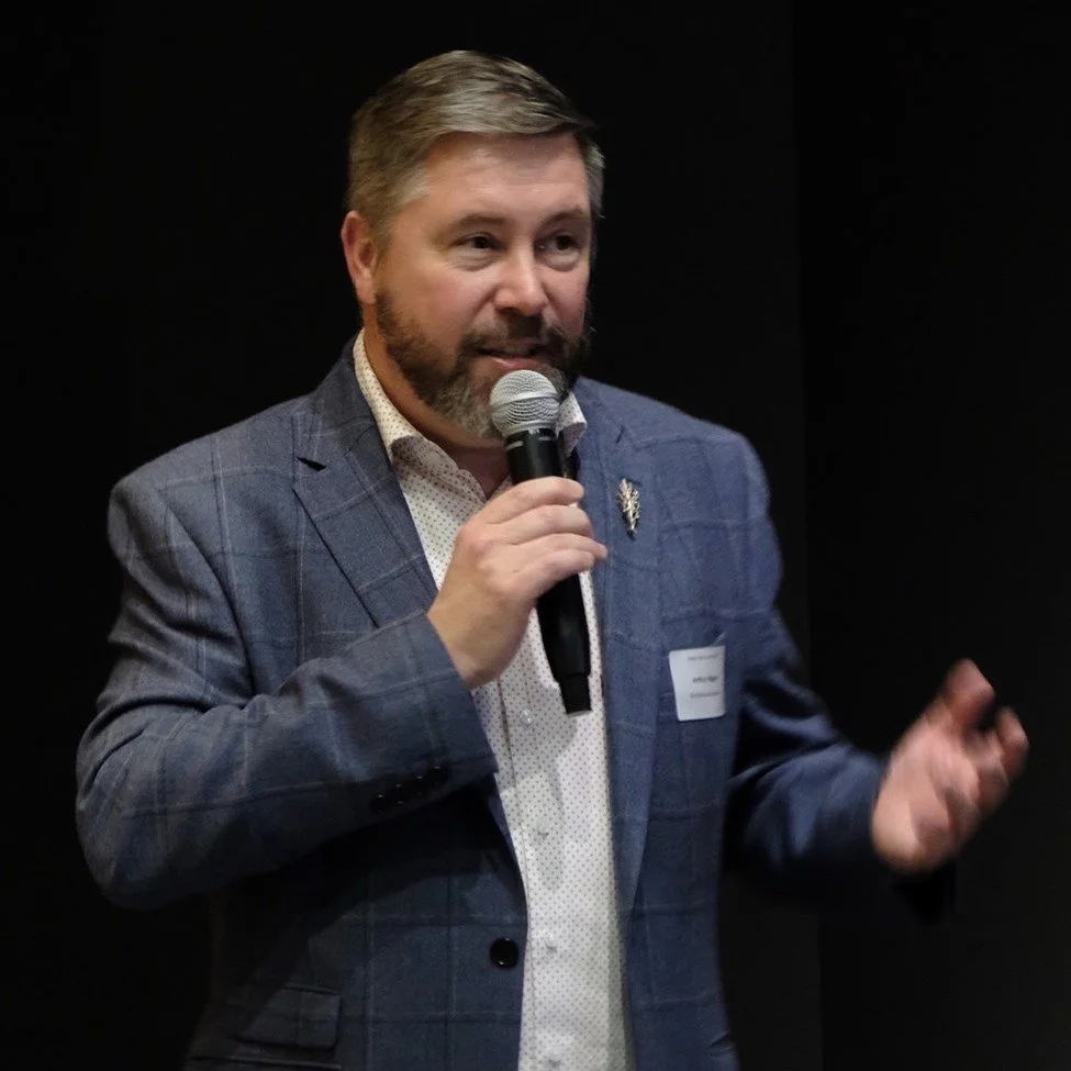 A man with light brown hair and a beard speaking into a microphone at an event, wearing a checked blue blazer and a white shirt with small patterns, with a dark background.