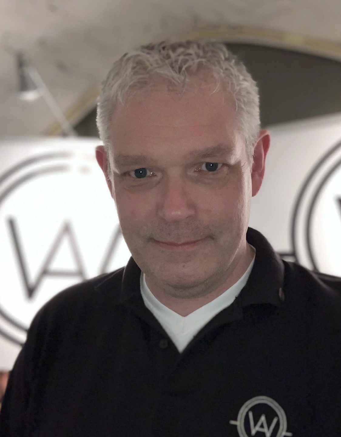 A man with short gray hair and blue eyes wearing a black shirt with a white undershirt, standing in front of a lighted sign with the letters 'W' and 'A'.