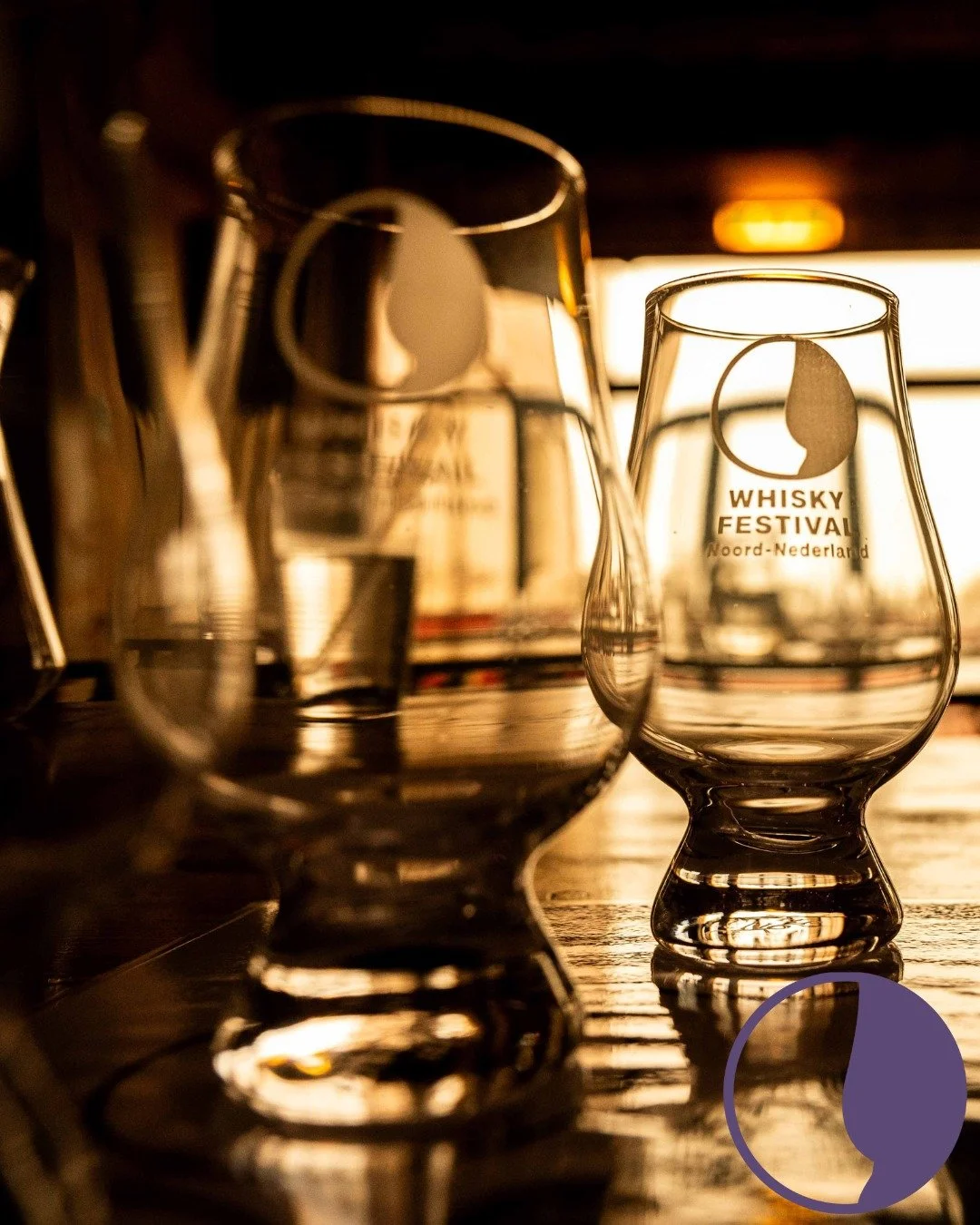 Two whisky glasses, one with the logo 'Whisky Festival Noord-Nederland', on a wooden surface with a warm backlight.