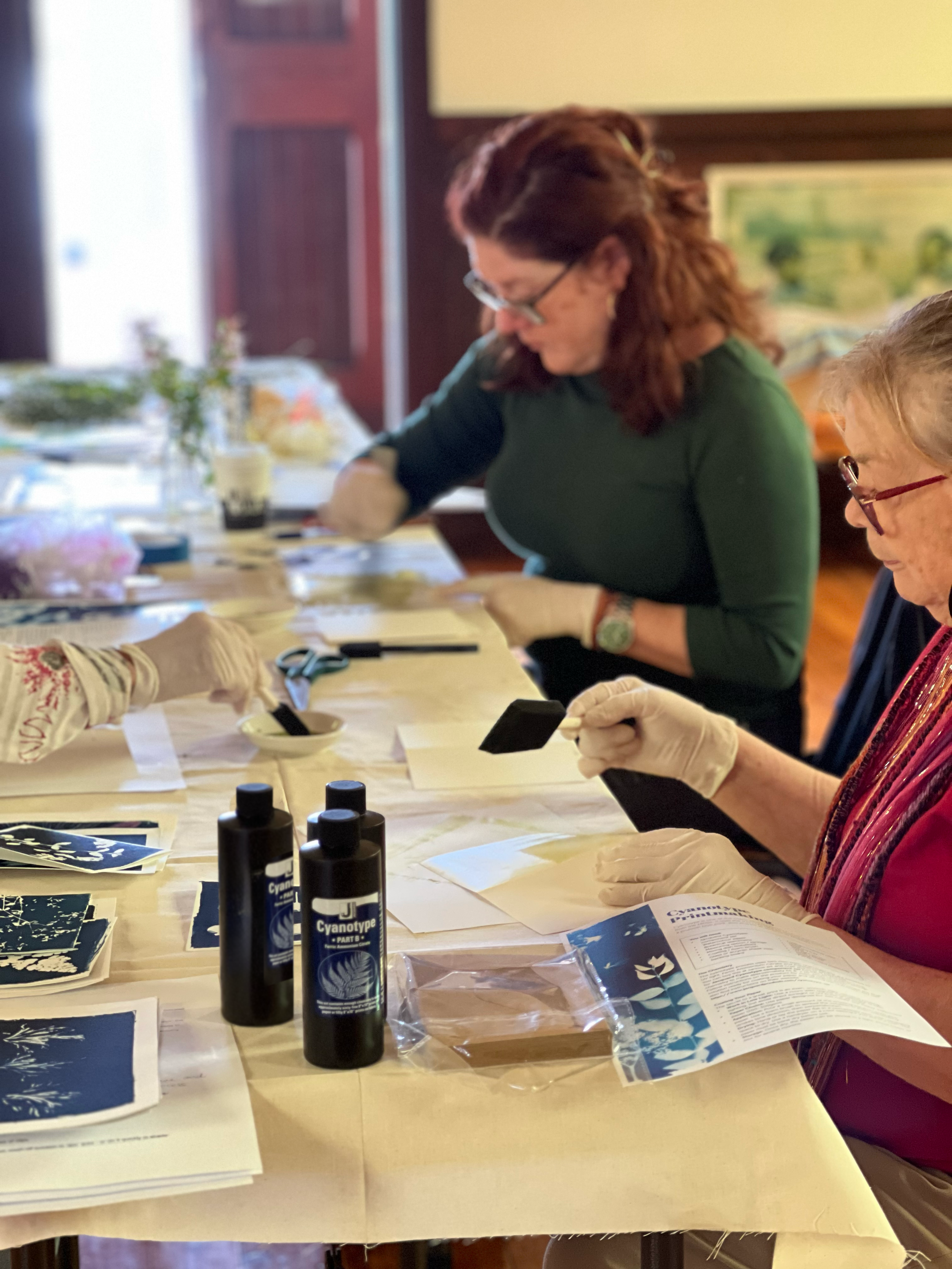 Two women working on cyanotype prints at a table, wearing gloves and examining the images with cyanotype chemicals bottles and papers around them.