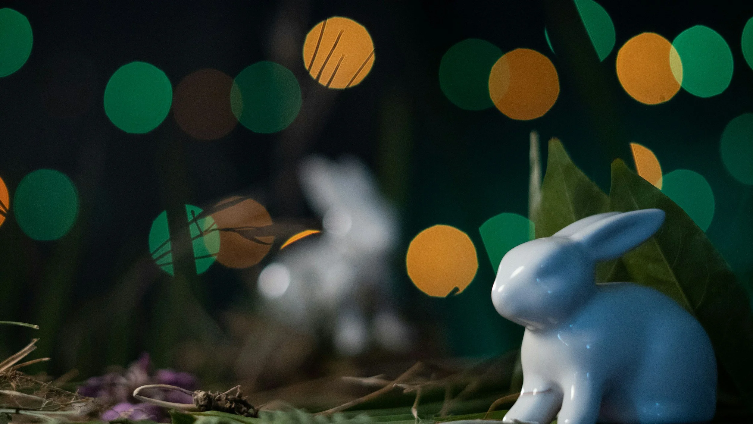 A white ceramic rabbit figurine sitting among dried leaves and purple flowers, with green leaves nearby, against a background of multicolored bokeh lights.