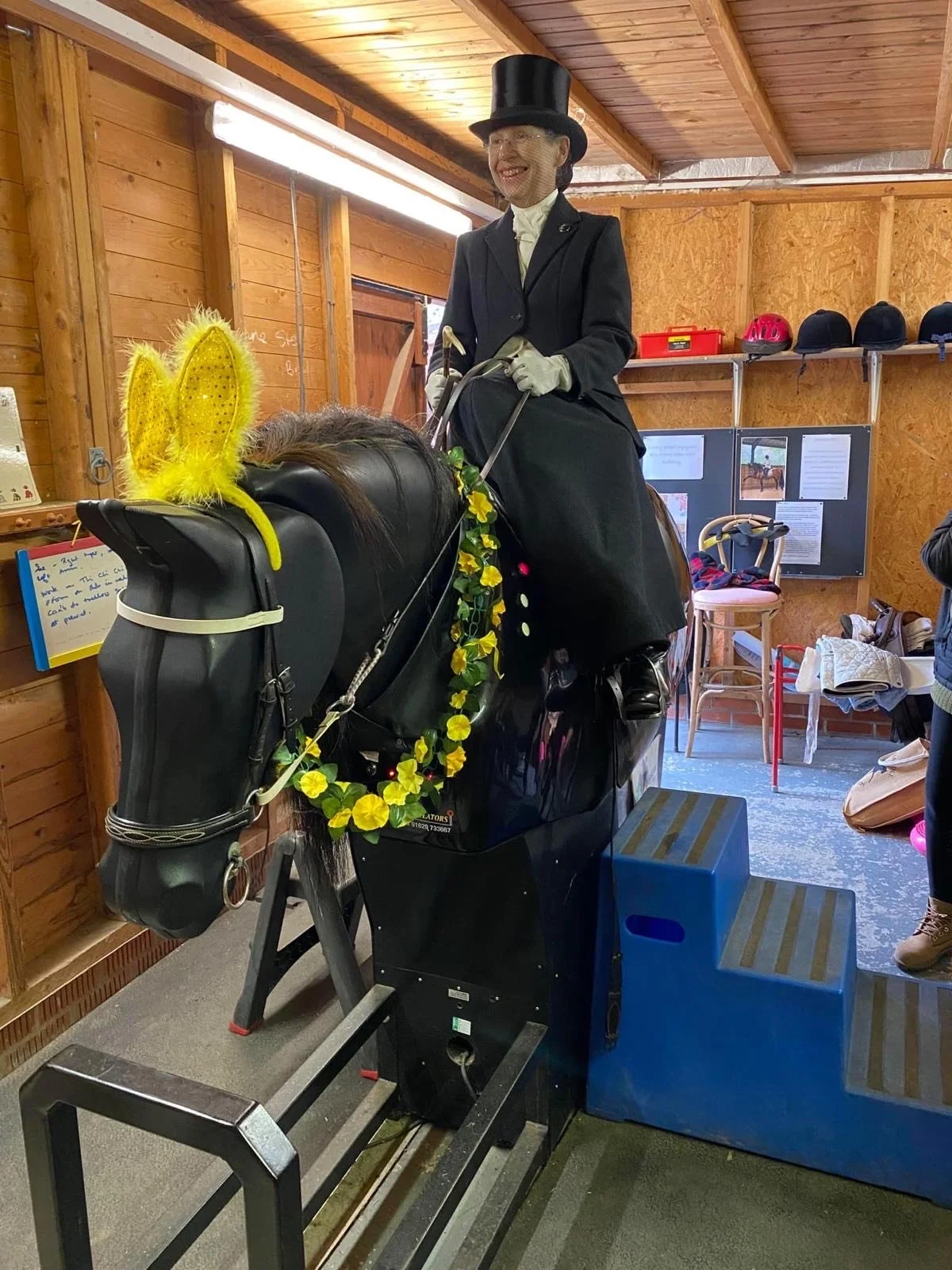 A woman dressed as a horse rider in a formal riding outfit with a top hat riding a mechanical horse in a wooden indoor space.