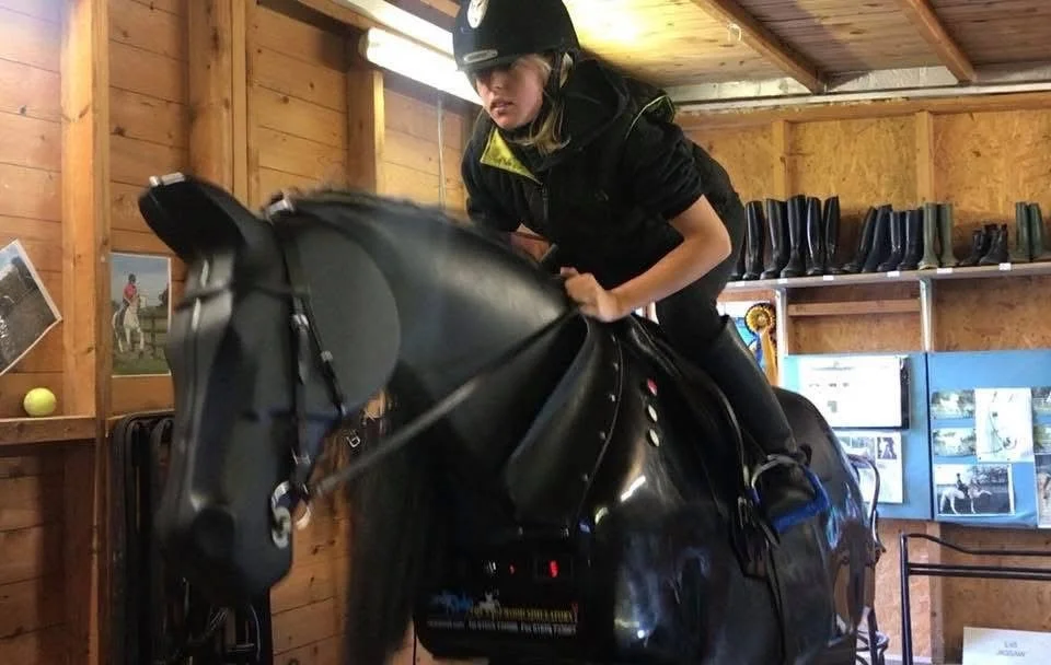 A person wearing a helmet and riding gear is riding a mechanical horse in an indoor stable area.