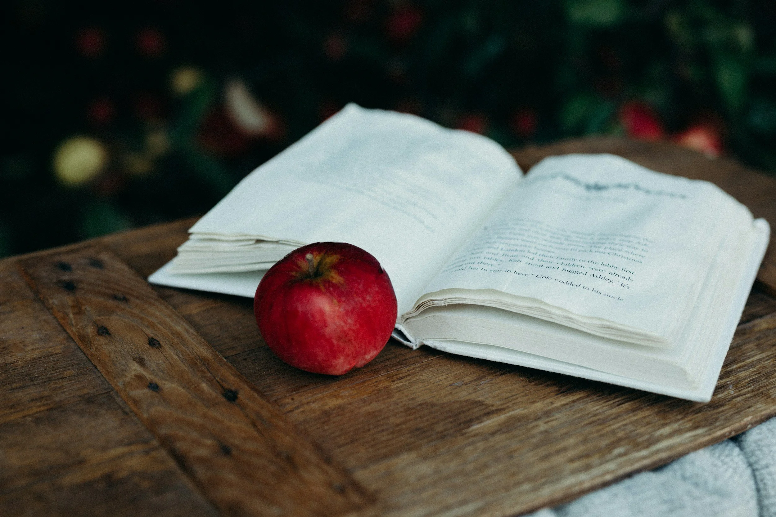 Open book and red apple on wooden surface, with blurred green foliage and red flowers in background.