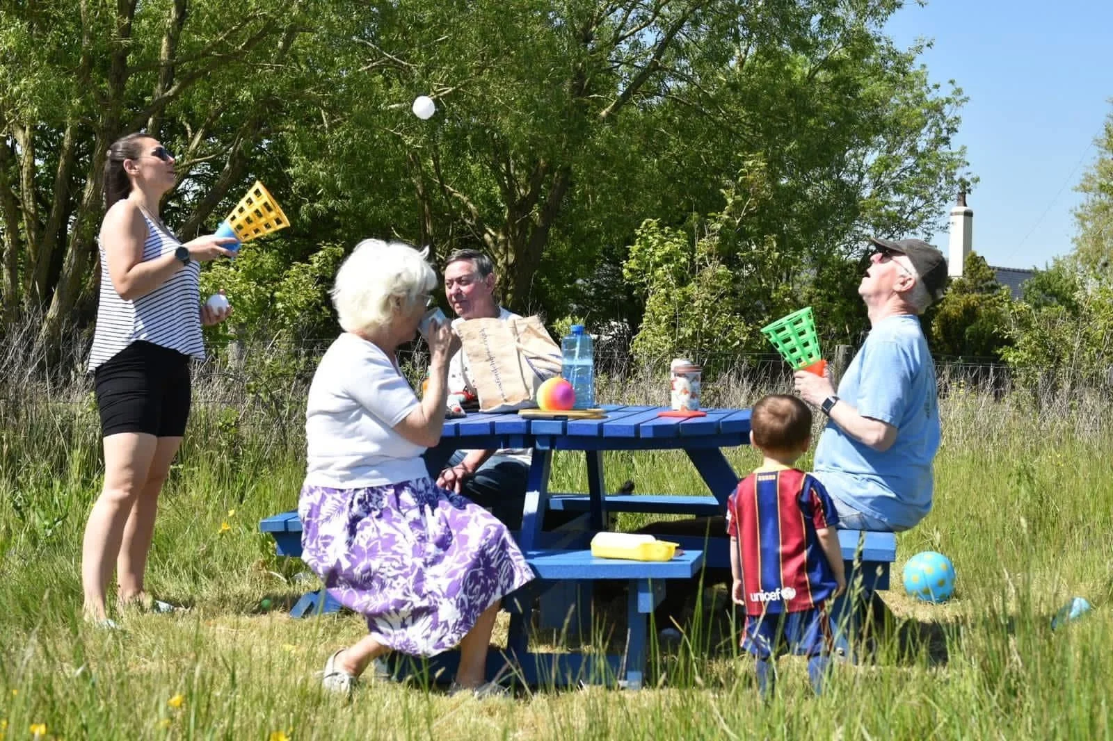 Group of people playing a game with balls and plastic cups outdoors on a sunny day, sitting and standing around a blue picnic table in a grassy area with trees.