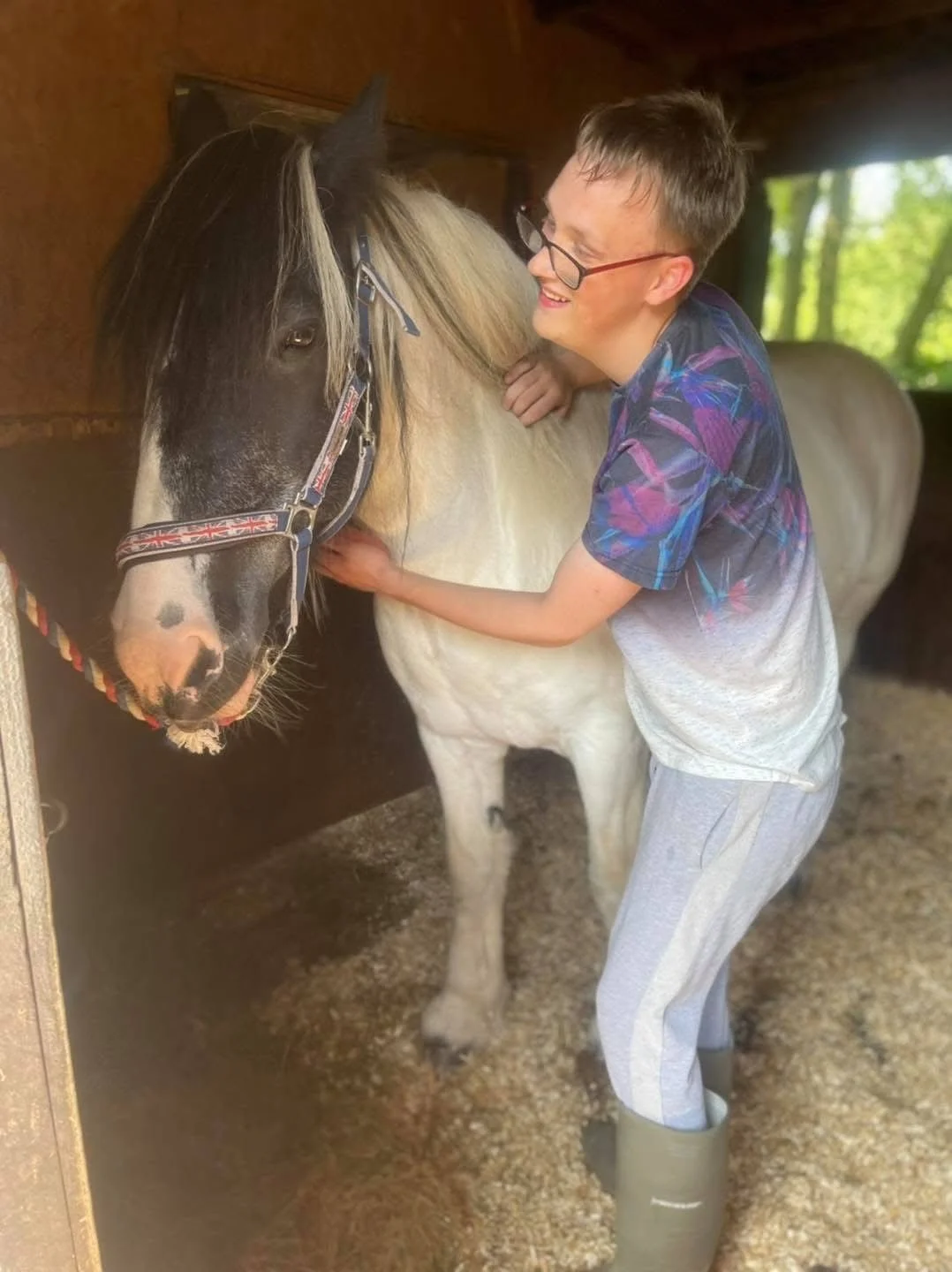 A young boy smiling and hugging a black and white horse inside a stable with green trees visible outside.