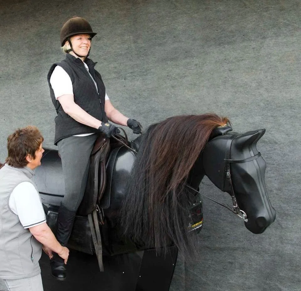 A woman wearing a helmet sitting on a black horse simulator with a person standing next to her, in front of a gray background.