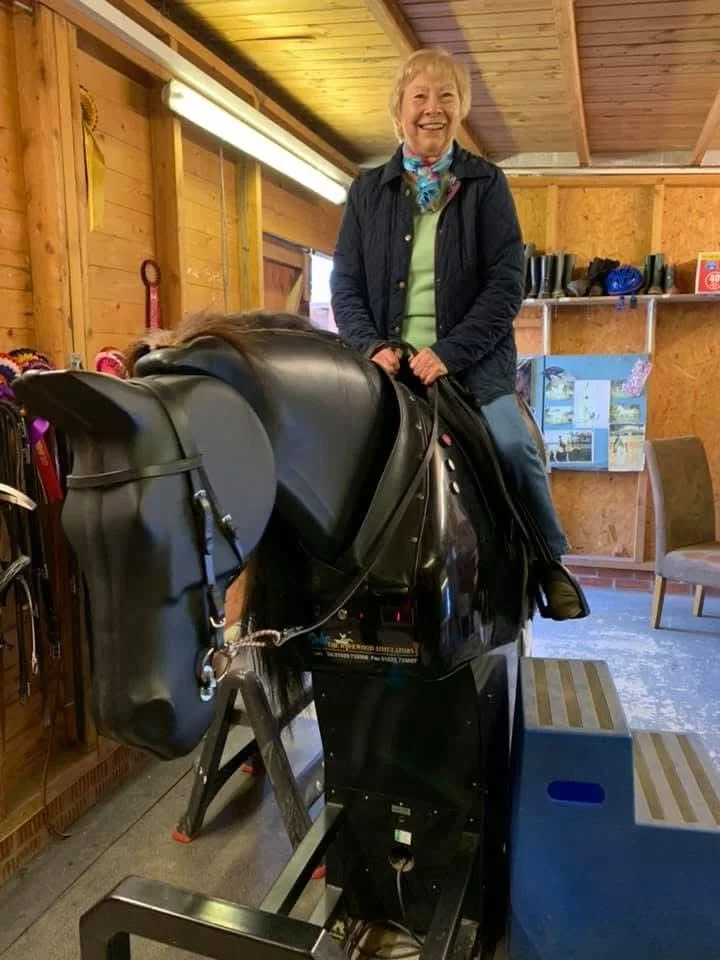 A smiling elderly woman sits on a mechanical horse inside a wooden stable.