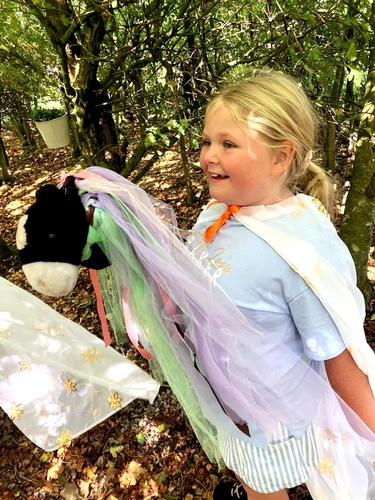 A young girl in a white t-shirt and striped shorts, with a colorful tulle cape, smiling in a wooded area, holding a stuffed horse toy with a black and white face and a multicolored mane.