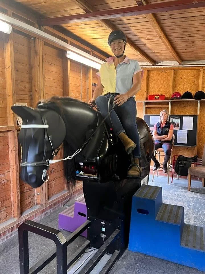 A woman smiling while riding a simulated horse in an indoor riding simulator, with another woman sitting in the background watching.