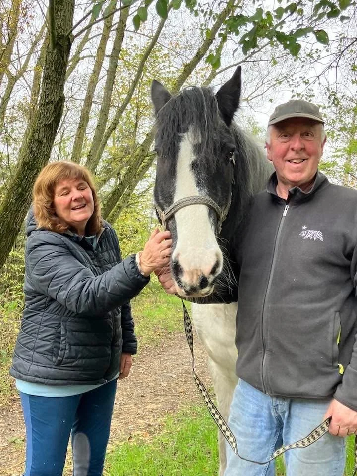 A woman and a man standing outdoors in a wooded area, smiling at the camera, holding a black and white horse with a halter. The woman is on the left, and the man is on the right.