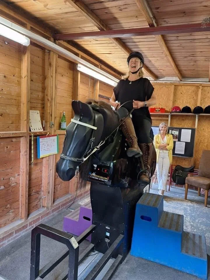 Woman laughing while riding a mechanical horse simulator in a wooden indoor space, with another woman watching in the background.