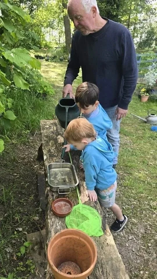 An elderly man with two young boys gathered around a small outdoor water activity station with a makeshift wooden water channel, funnels, and containers in a garden setting.