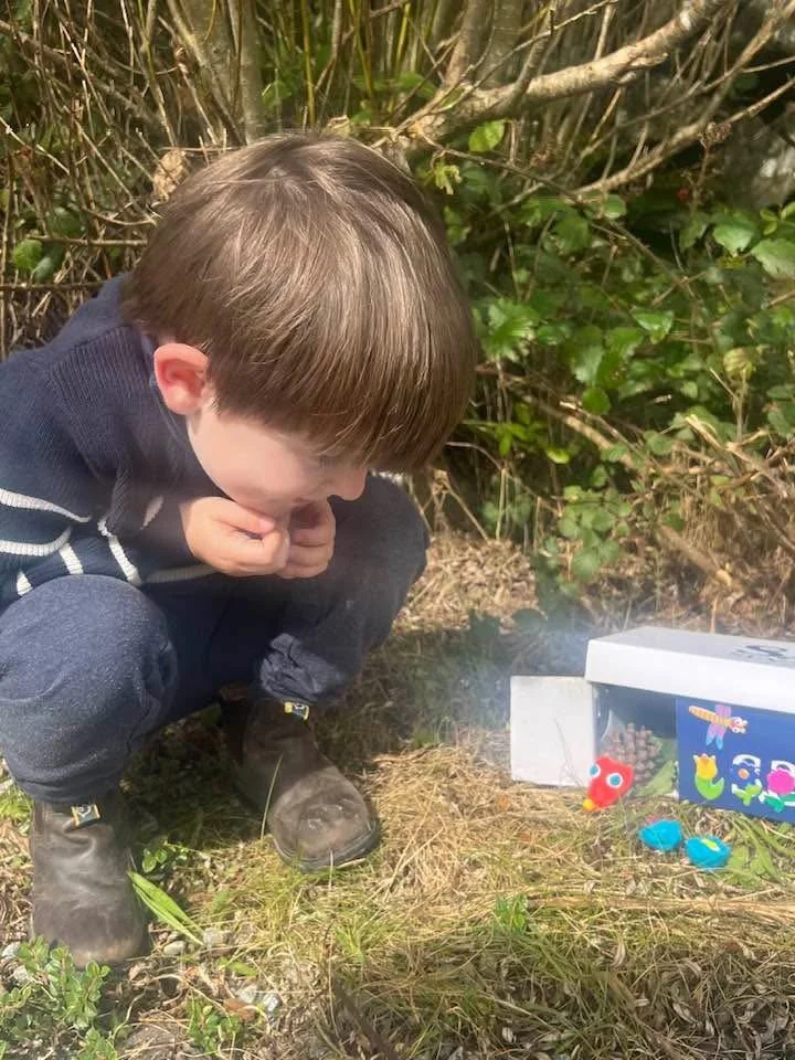 Young boy crouching outdoors, intently observing a small, colorful toy bird and other toys on the ground next to a box of similar toys, surrounded by bushes and grass.