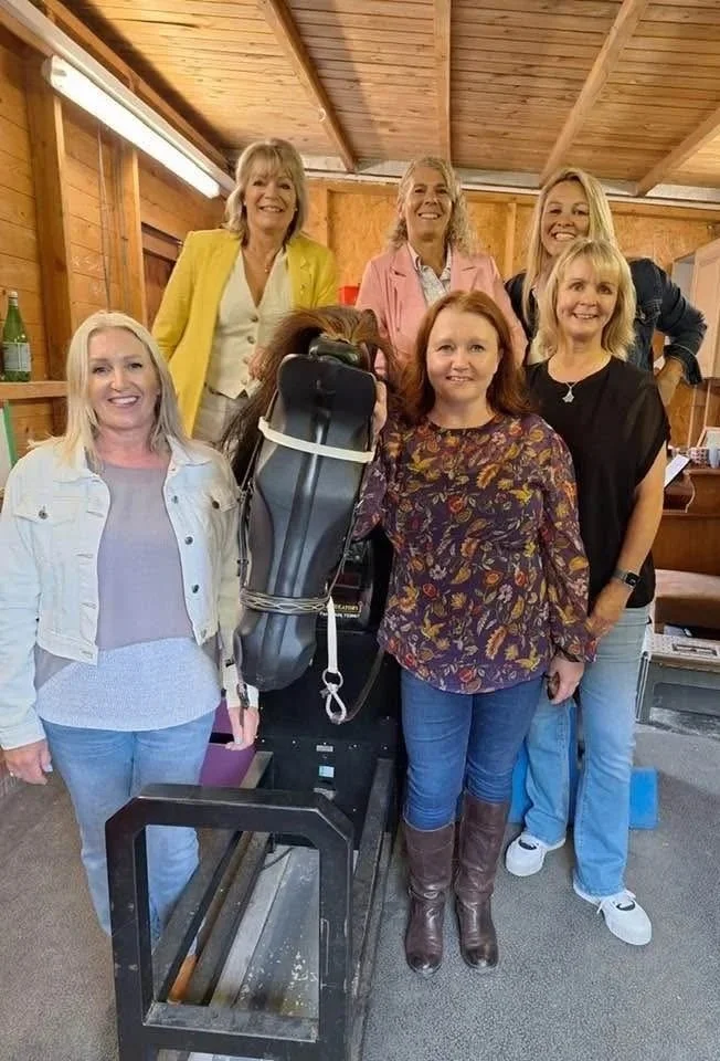 Group of six women standing in a wooden room, smiling for a photo. One woman is wearing a yellow blazer, another in a pink blazer, and others in casual tops. They are gathered around a large piece of equipment.
