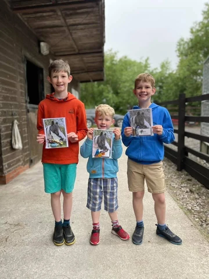 Three boys holding up pictures of a horse with a white background, standing outdoors on a concrete surface near a wooden building with a black fence and green trees in the background.