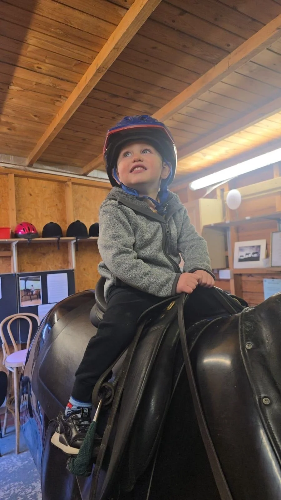 A young boy sitting on a black horse riding simulator, wearing a helmet, inside a wooden room, with shelves and framed pictures in the background.