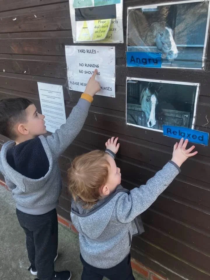 Two children looking at photos of horses on a wooden wall; one photo shows a horse with an angry label, and the other shows a relaxed horse. There are yard rules posted above.