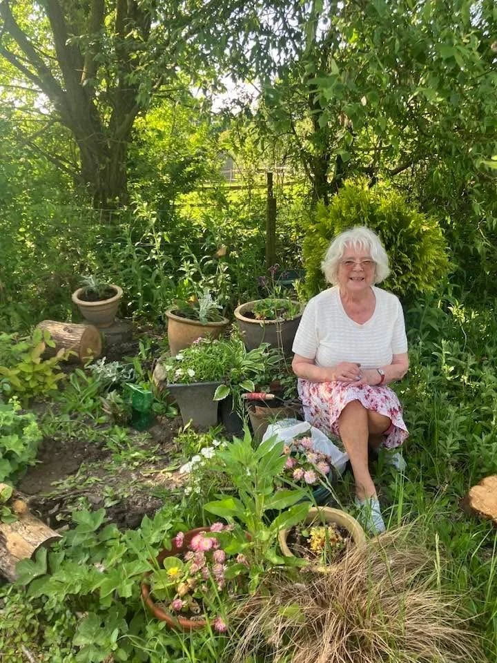 An elderly woman with white hair smiling and sitting outdoors in a garden with various plants and potted flowers.
