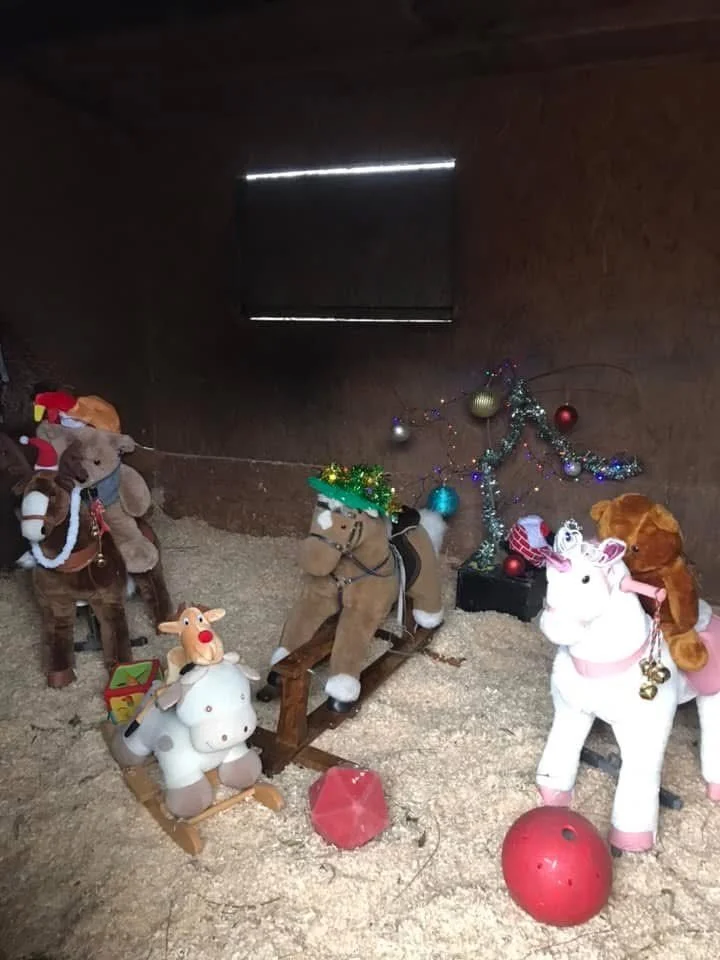 Festive Christmas decorations with plush toy reindeer and unicorns, a small Christmas tree with ornaments, and lights in a wooden room with a small window at the back.
