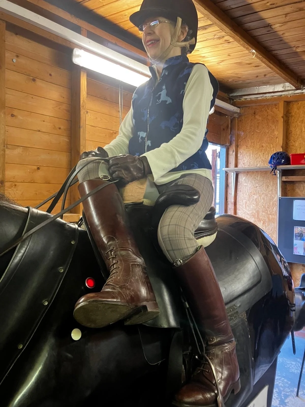 A woman riding a horse simulator housed in an indoor wooden stable, wearing a black helmet, glasses, a blue vest with horse patterns, plaid riding pants, and tall brown riding boots.