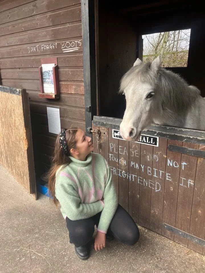 A girl kneeling in front of a horse stall, looking up at a white horse looking out from the stall.