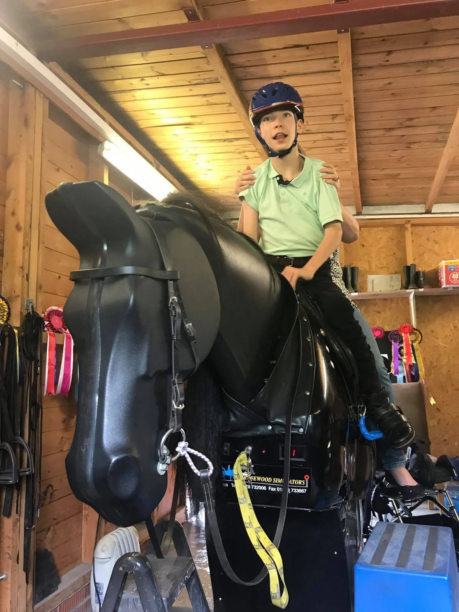 Child wearing a blue safety helmet riding a black mechanical horse simulator with instructor hands on kid's shoulders inside a wooden stable or riding arena.