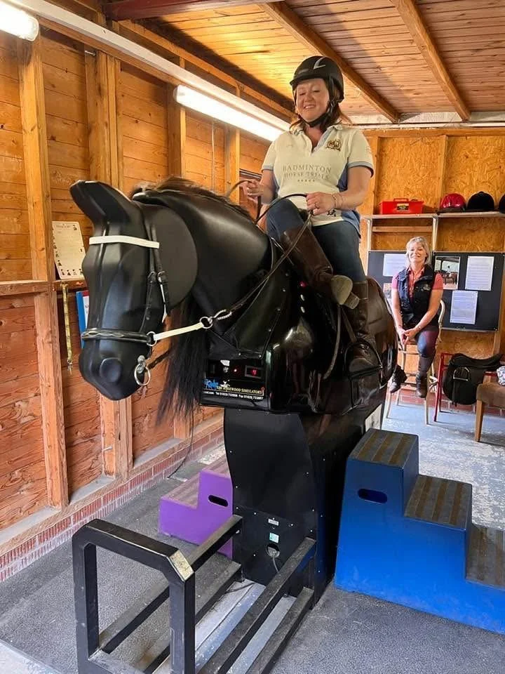 A woman in a riding helmet and shirt is riding a mechanical horse simulator in a wooden room. The machine has blue and purple sections at its base for support, and a woman is sitting in the background smiling.