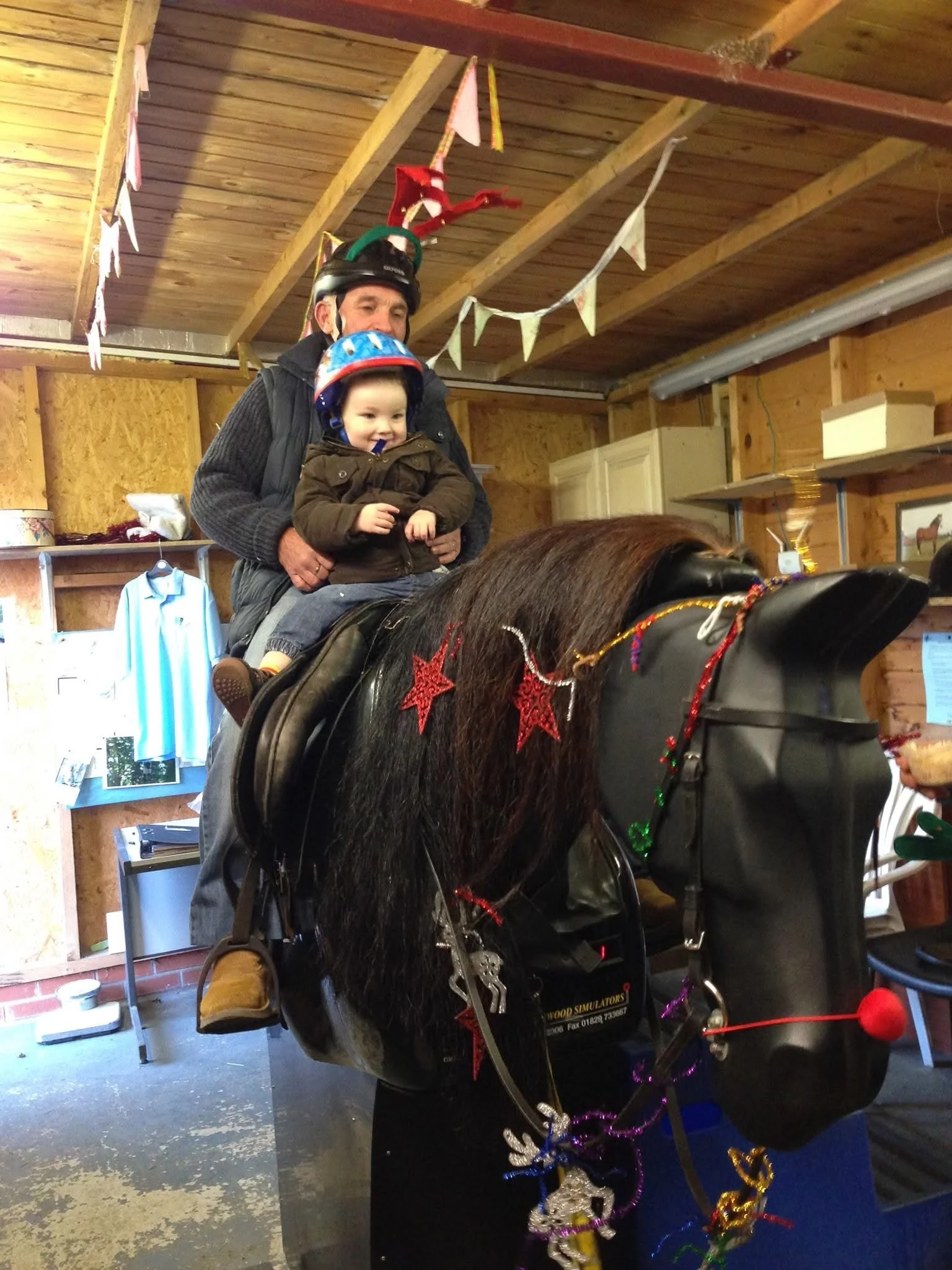 A man and a young child riding a mechanical horse at an indoor event, decorated with Christmas ornaments and garlands, under a wooden ceiling with festive banners.