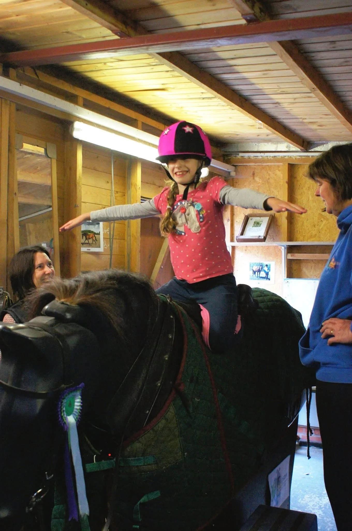 A young girl wearing a pink helmet and a pink and gray shirt is sitting on a horse indoors, with her arms outstretched and a big smile. Two women are standing nearby, watching her.