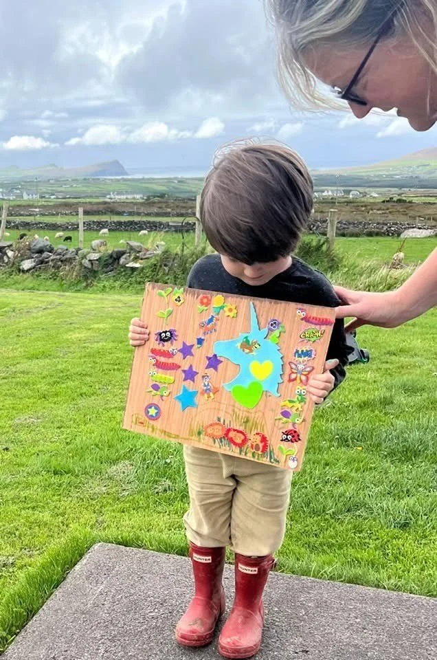 A young boy holding a colorful foam board with stickers of sea creatures, stars, and cartoon characters, standing outdoors on a patch of concrete with a grassy landscape and cloudy sky in the background. An adult woman is leaning over to look at the board.
