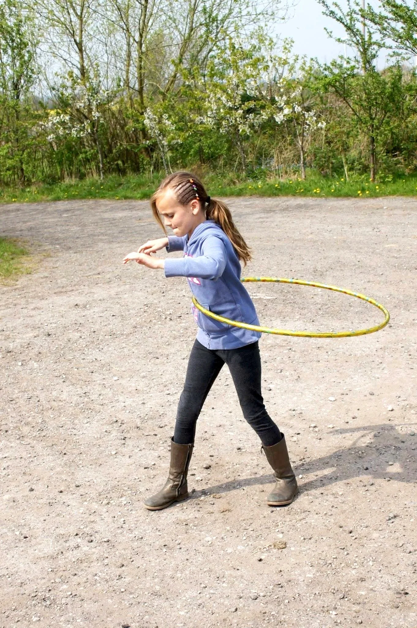 A girl playing with a hula hoop outdoors on a dirt path with trees and bushes in the background.