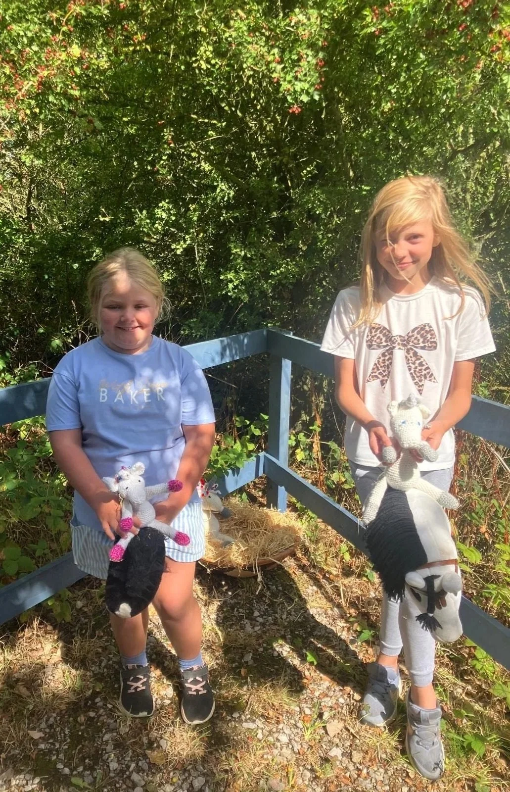 Two young girls standing outdoors in a garden area with greenery and a blue railing, each holding a knitted stuffed animal horse, smiling at the camera on a sunny day.