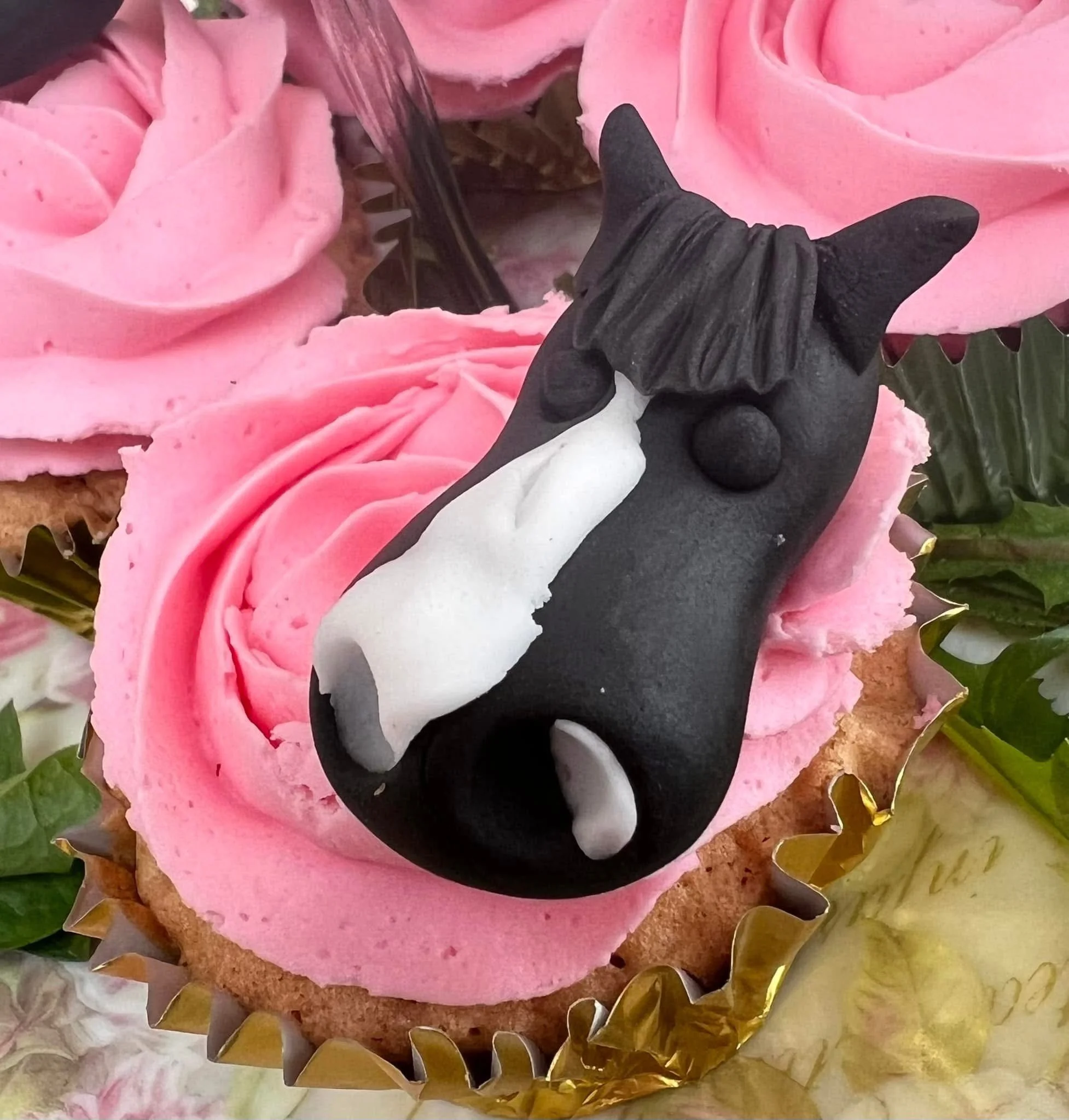 Pink frosted cupcake topped with a black horse-shaped cookie with a white blaze and a chocolate ball nose, surrounded by pink roses and green leaves.