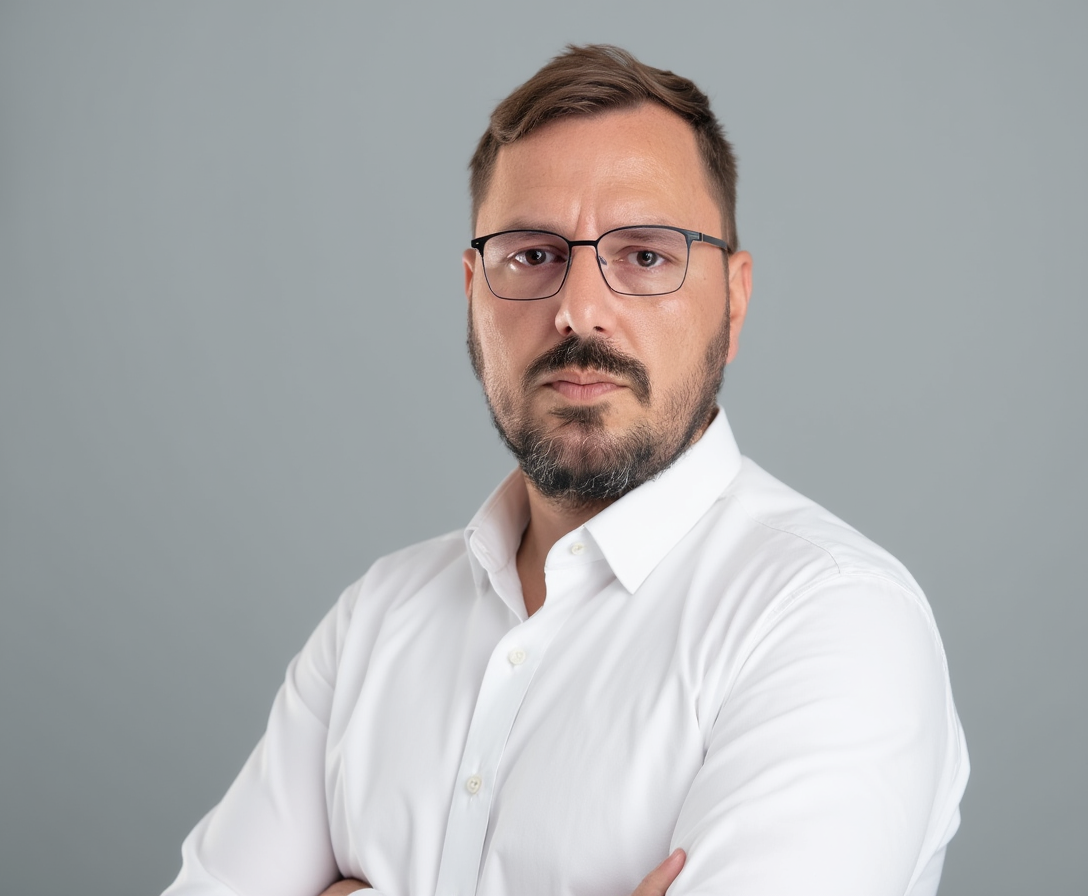 Ein ernst blickender Mann mit Brille, kurzen Haaren und Bart trägt ein weißes Hemd und steht vor einem grauen Hintergrund.